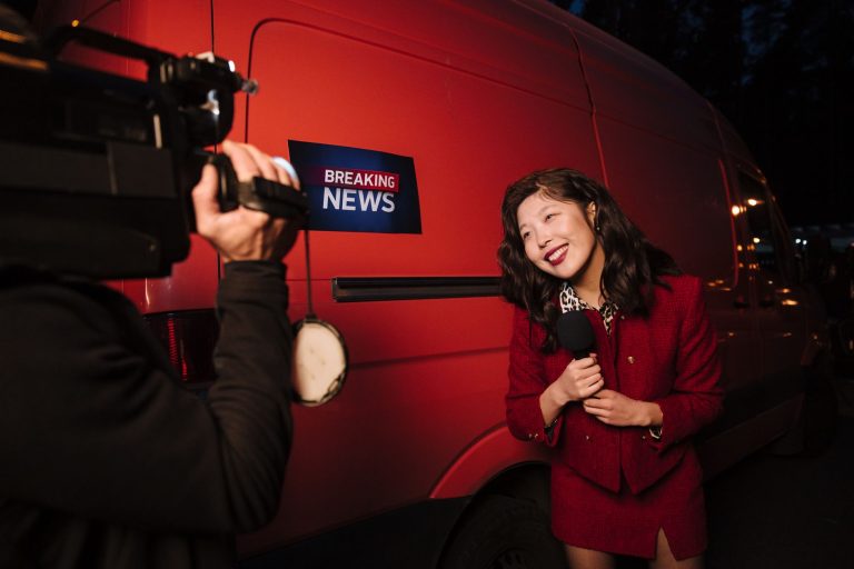 Asian female reporter in a red blazer reporting live on the news outdoors in the evening. — TonySpins Casino und seine innova